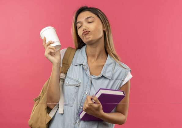 女孩可爱的年轻漂亮的女学生戴着背袋拿着书和塑料咖啡杯闭着眼睛孤立在粉红色的墙上包杯子粉色