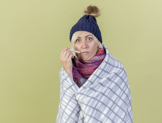 人给人印象深刻的年轻金发病女人戴着冬天的帽子和围巾包裹在格子布把温度计放在嘴里隔离在橄榄绿的墙上嘴姿势金发