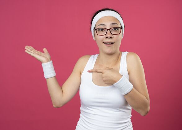 眼镜戴着眼镜戴着头带和腕带的年轻运动女性惊讶地指着隔离在粉红色墙上的空手点年轻惊喜