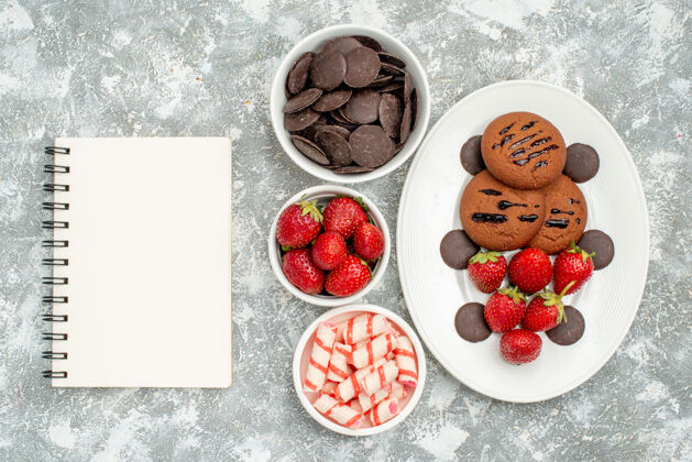 碗俯瞰图：白色椭圆形盘子上的巧克力饼干草莓和圆形巧克力 灰白色地面上的碗和糖果草莓巧克力和笔记本草莓甜点糖果