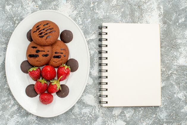 草莓顶部近距离观看巧克力饼干草莓和圆形巧克力在白色椭圆形盘子在左边和一个笔记本在灰白色的地面右侧盘子椭圆形覆盆子