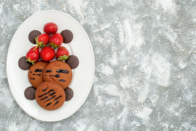 左边左上角是灰白色地面上白色椭圆形盘子上的巧克力饼干草莓和圆形巧克力早餐蓝莓水果