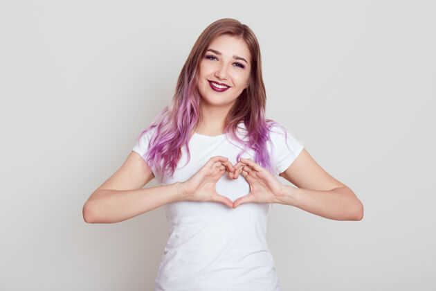心快乐积极的女性 明亮的淡紫色头发 迷人的微笑 用手指表示心意 表达爱 在灰色的墙上摆出孤立的姿势情感表达女性