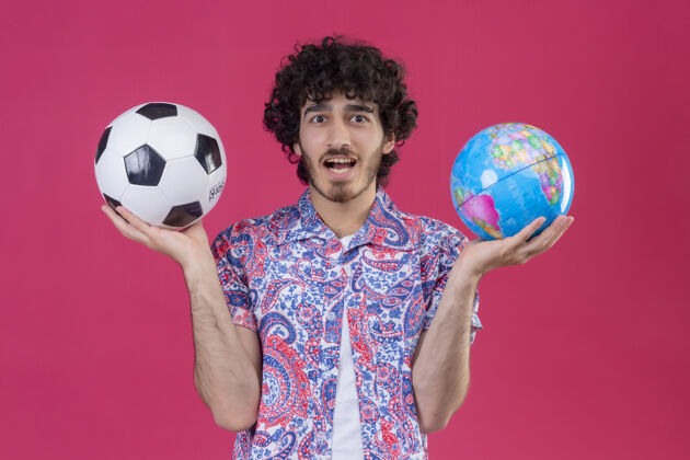 印象令人印象深刻的年轻英俊卷发旅行者男子拿着地球仪和足球在孤立的粉红色空间地球仪旅行者年轻