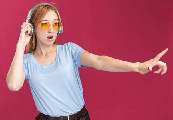 红发自信的年轻红发姜黄色女孩 戴着太阳镜和耳机 脸上长着雀斑 一边看着一边指着粉色耳机自信阳光