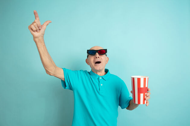 手指着电影院的眼镜蓝色工作室背景上的高加索老人肖像美丽的情感模型人类情感的概念 面部表情 销售 福利 广告空间成功面部衬衫
