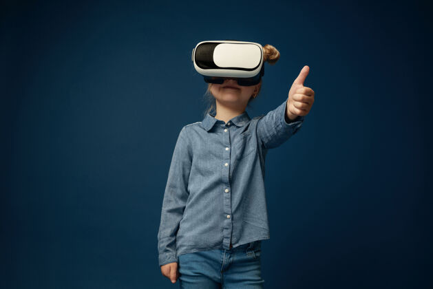 现实展示你的想法小女孩或孩子穿着牛仔裤和衬衫 戴着虚拟现实耳机眼镜 与蓝色工作室背景隔离尖端技术的概念 视频游戏 创新模拟操纵杆梦想