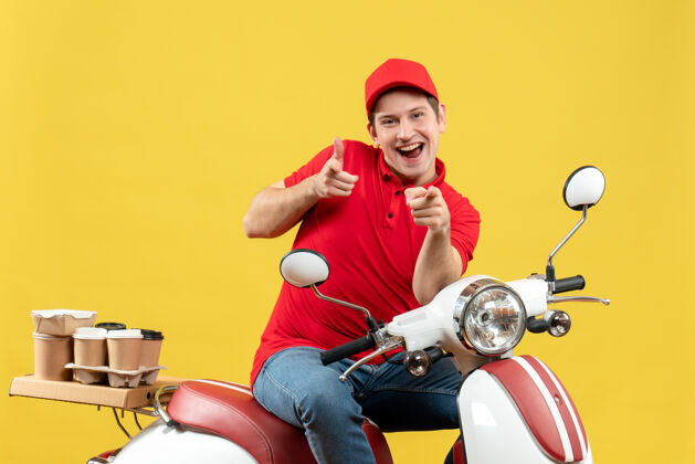 帽子俯视图：微笑的年轻人穿着红色上衣 戴着帽子 坐在滑板车上 指着黄色的墙上交付指向年轻人