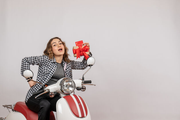音乐前视图可爱的年轻女子骑着轻便摩托车拿着礼物和卡片在灰色的墙上举行前面快乐