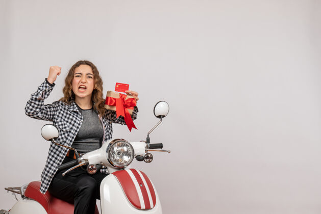 肖像前视图愤怒的年轻女子骑着轻便摩托车拿着礼物在灰色的墙上快乐摩托车礼物