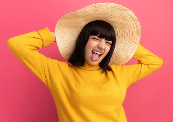 黑发快乐的年轻黑发白人女孩戴着沙滩帽 伸出舌头 把头放在粉红色的衣服上快乐舌头年轻