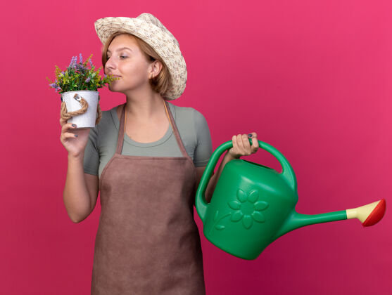 花园高兴的年轻斯拉夫女园丁戴着园艺帽拿着浇水罐 嗅着粉红色花盆里的花园艺女性帽子