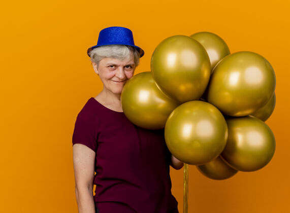 帽子高兴的老妇人戴着聚会帽 手里拿着氦气球 隔离在橙色的墙上 留着复制空间氦气气球拿着