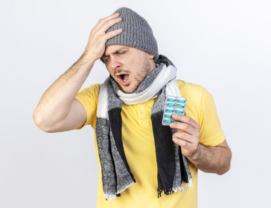 抱疼痛的年轻金发病斯拉夫男子戴着冬季帽子和围巾 手放在头上 拿着一包药片 隔离在白色墙壁上 留有复印空间痛疾病穿