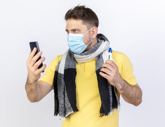 金发令人印象深刻的年轻金发病斯拉夫男子戴着医用口罩和围巾看着电话 拿着温度计隔离在白色墙上的复制空间电话疾病持有