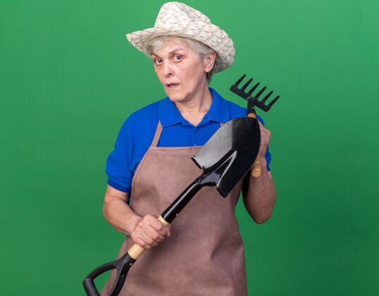 花园焦虑的上了年纪的女园丁戴着园艺帽拿着铲子和耙子在草地上老人女园艺