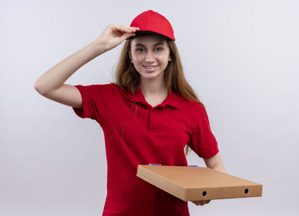 帽子微笑着的年轻送货女孩 穿着红色制服 拿着包裹 把手放在隔离的空白处的帽子上年轻交货举行
