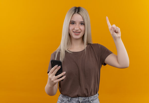 年轻戴着牙套微笑的金发女孩举着手机 手指放在橙色的空白处 还有复印空间金发太空复制