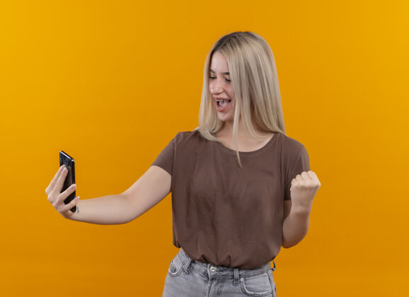 提高快乐的年轻金发女孩戴着牙套拿着手机看着它举起拳头在孤立的橙色空间与复制空间持有电话橙色