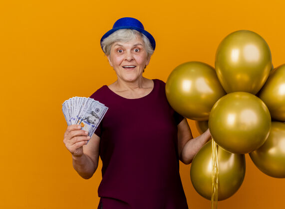 钱惊讶的老妇人戴着聚会帽 手里拿着氦气球和钱 隔离在橙色的墙上 还有复印空间惊喜穿女人