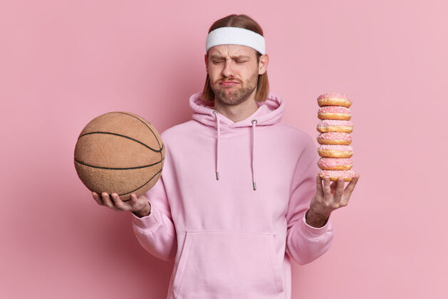 玩人们运动垃圾食品的概念不高兴的运动型男子看着篮球不高兴持有一堆甜甜的开胃甜甜圈不想玩穿连帽衫人运动休闲