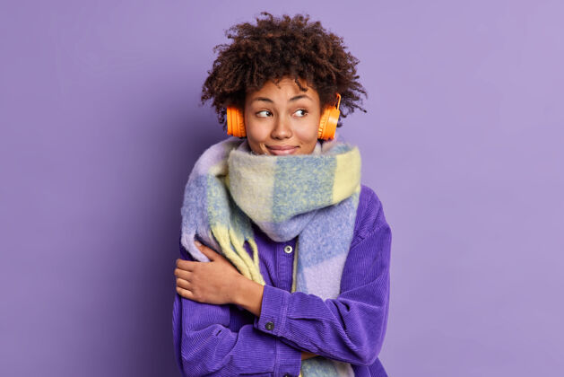 正面可爱体贴的非洲裔美国妇女拥抱自己温暖的脖子上戴着围巾 通过无线耳机听音乐时愉快地集中在一边使用快乐旁听