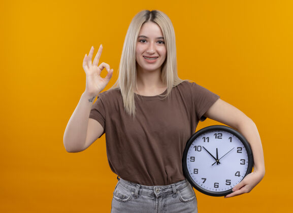 时钟微笑着的金发女孩戴着牙套拿着时钟在孤立的橙色空间和复印空间做ok标志好年轻牙套