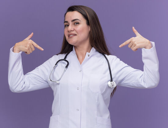 听诊器高兴的年轻女医生穿着医用长袍 用听诊器指着自己 双手放在紫色上请年轻长袍