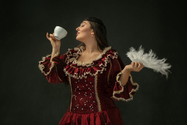 农民喝着咖啡 捧着蓬松的扇子中世纪的年轻女子穿着红色复古服装 背景是深色的女模特是公爵夫人 皇室的人观念比较时代 现代 时尚 美丽皇后好公爵夫人