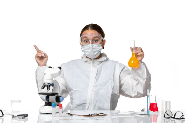 指着正面图：女医生穿着特制的工作服 戴着口罩 拿着装有黄色溶液的烧瓶 背景是白色的冠状病毒大流行病毒套装外套白色