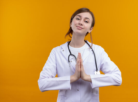 听诊器高兴的年轻女医生穿着医疗袍和听诊器做祈祷手势上孤立的橙色空间与复制空间橙色长袍穿