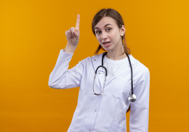 年轻印象深刻的年轻女医生穿着医用长袍和听诊器举起手指在孤立的橙色空间与复制空间穿着提高太空
