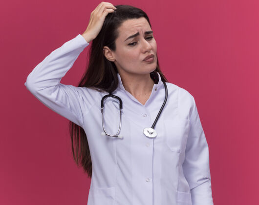 听诊器困惑的年轻女医生穿着带听诊器的医用长袍 手放在头上 低头看着粉红色女医学手