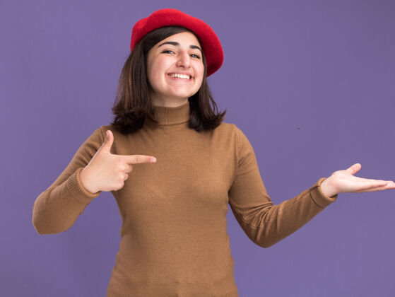 帽子微笑着的年轻漂亮的白人女孩戴着贝雷帽指着紫色的空手指向贝雷帽漂亮