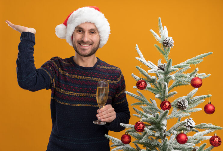 男人快乐的白人年轻人戴着圣诞帽 站在装饰好的圣诞树旁 手里拿着一杯香槟 看着摄像机 在橙色背景下 一只空手孤立着圣诞站着年轻