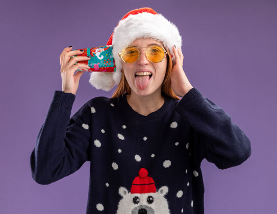 帽子快乐的年轻漂亮女孩 穿着圣诞毛衣 戴着圣诞帽 戴着圣诞眼镜 耳朵上戴着圣诞杯 舌头孤立在紫色的背景上表演圣诞节舌头
