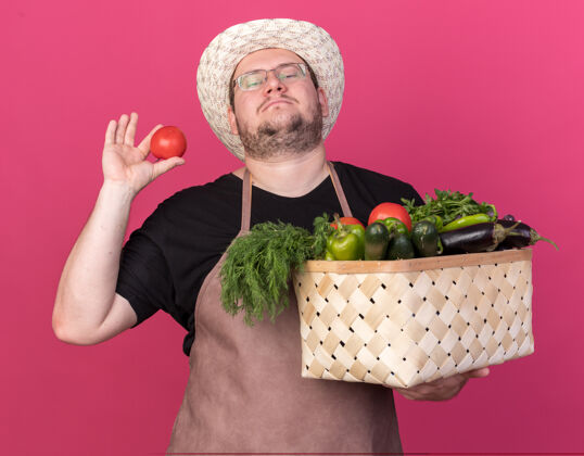 帽子自信的年轻男园丁戴着园艺帽 手里拿着菜篮子和西红柿 隔离在粉红色的墙上园艺人姿势