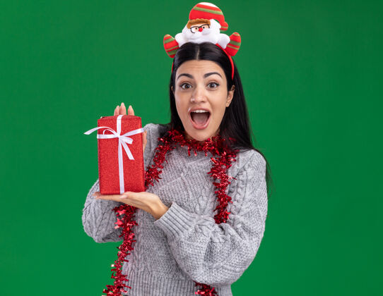 复制令人印象深刻的年轻白人女孩戴着圣诞老人的头带和镀金花环在脖子上拿着礼品包看着相机隔离在绿色的背景女孩周围圣诞快乐