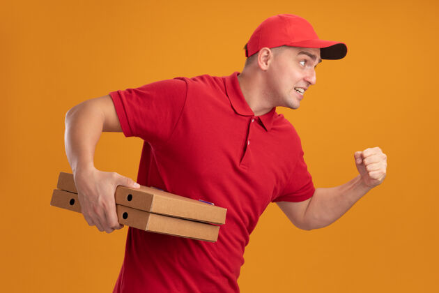 跑步紧张的年轻送货员身穿制服 头戴鸭舌帽 拿着披萨盒 在橙色的墙上孤立地展示着跑步的姿势年轻人男人拿着