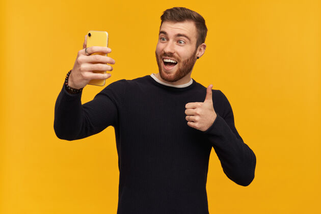 手机留着胡子的兴奋的 快乐的 深色头发的男人穿黑色毛衣自拍 竖起大拇指 表示赞同孤立地站在黄色的墙上外表颜色年轻