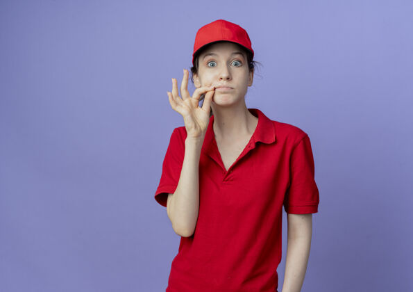 印象令人印象深刻的年轻漂亮的送货女孩穿着红色制服 戴着帽子 看着相机拉链她的嘴关闭在紫色背景与复制空间隔离拉链制服背景