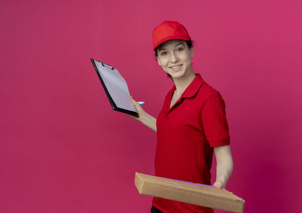 背景微笑着的年轻漂亮的送货女孩 穿着红色制服 戴着帽子 手里拿着披萨包和剪贴板 笔被隔离在深红色的背景上 还有复印空间披萨深红色女孩