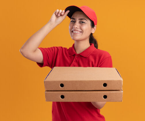 站着微笑的年轻送货女孩穿着制服 戴着帽子 拿着披萨盒和帽子 隔离在橙色的墙上衣服年轻人脸