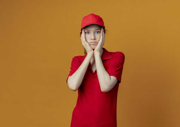 橙色困惑的年轻漂亮的送货女孩 穿着红色制服 戴着帽子 手放在脸上 看着隔离在橙色背景和复印空间的相机帽子女孩制服