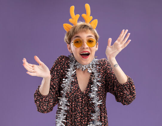 年轻兴奋的年轻漂亮女孩戴着驯鹿鹿角头带 脖子上戴着金箔花环 戴着眼镜 看着镜头 紫色背景上孤立的空手圣诞快乐鹿角花环