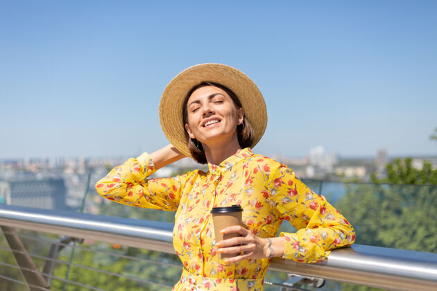 年轻人穿着黄色夏装 戴着黄色帽子 喝着咖啡 享受阳光的女人的户外肖像 站在桥上 城市的景色令人惊叹欢乐成人深色头发