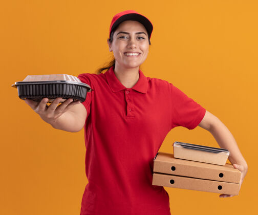 女孩微笑着的年轻送货女孩穿着制服 戴着帽子 拿着披萨盒 拿着食物容器站在橙色的墙上人抱着微笑