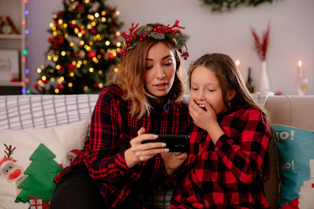 享受令人印象深刻的母亲和女儿坐在沙发上看电话 享受圣诞节的时间在家里印象圣诞快乐圣诞节