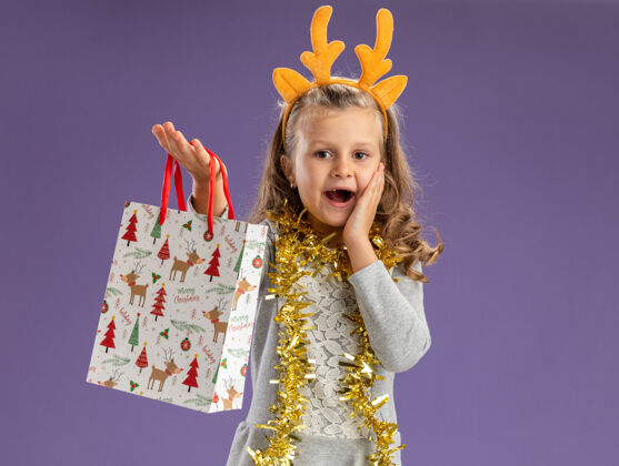 包兴奋的小女孩戴着圣诞发箍 脖子上戴着花环 手放在脸颊上 手放在蓝色背景上 向镜头伸出礼品袋礼物脖子抱