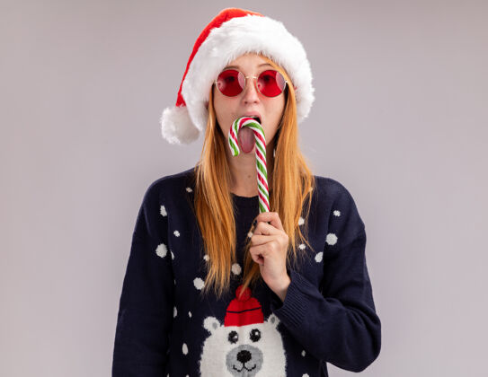 圣诞节看着镜头年轻漂亮的女孩戴着圣诞帽和眼镜拿着和舔着圣诞糖果隔离在白色背景上眼镜舔女孩